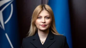 Жінка з довгим білявим волоссям у чорному піджаку на тлі прапорів НАТО